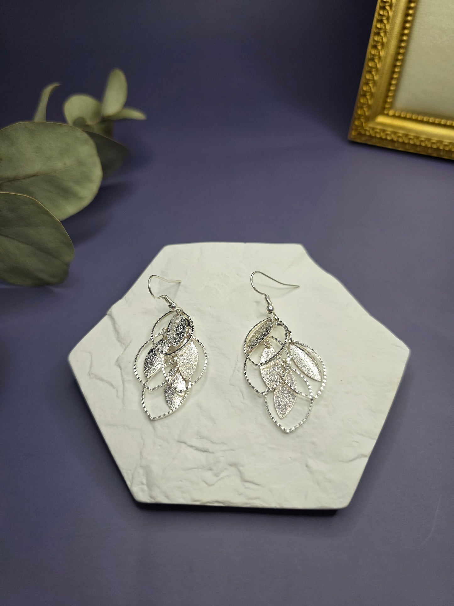 Glitzy Silver Filigree Leaf earrings displayed on a white textured hexagon stone. These 6cm botanical drop earrings feature a tiered design with a shimmering dusty texture and stainless steel ear needles - My Silver Lining.