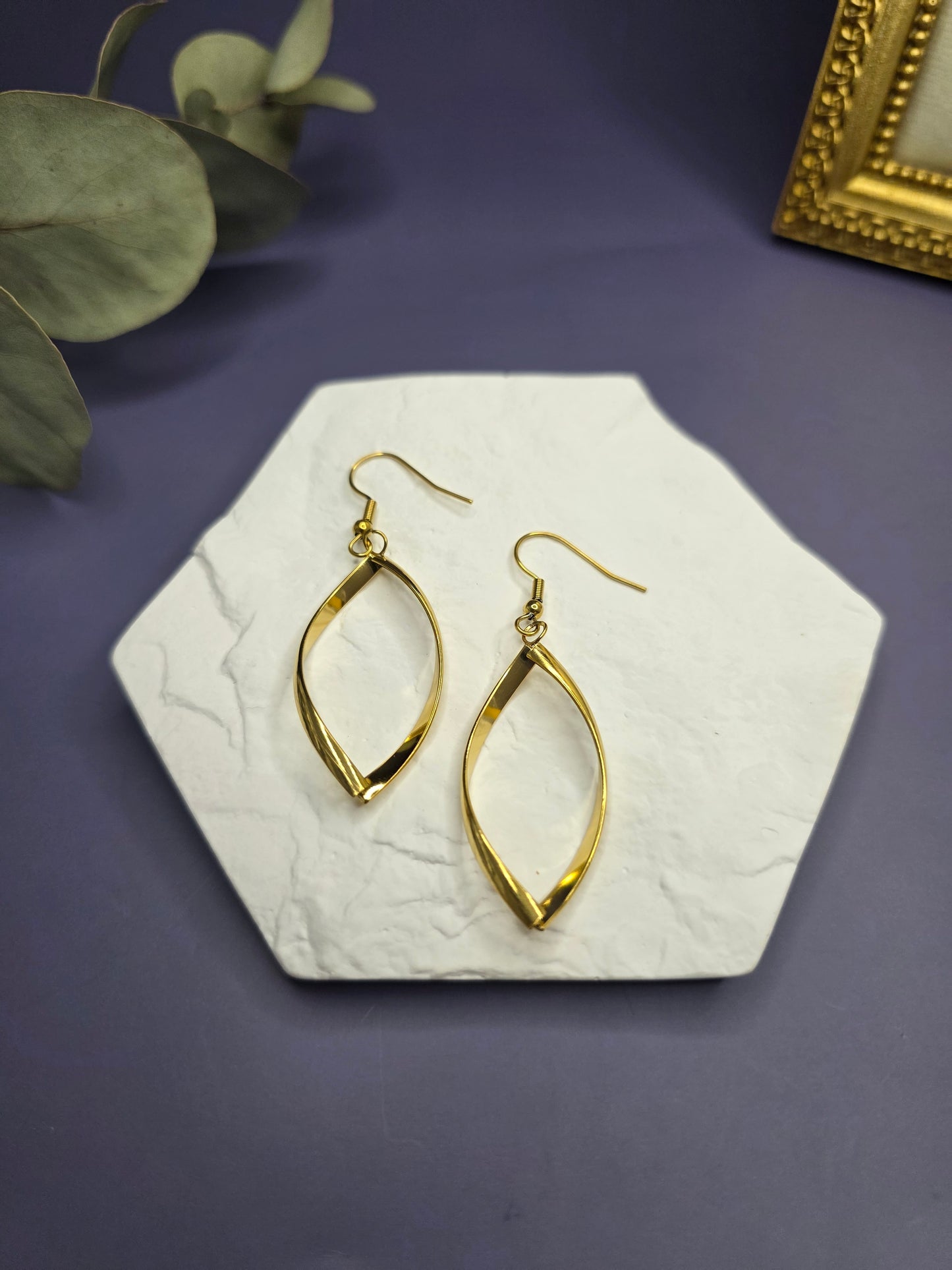 Giolden ribbon earrings displayed on a white textured hexagon stone. These 7cm waterproof 316 stainless steel drop earrings offer a polished, tarnish-resistant twisted design for modern everyday elegance - My Silver Lining.