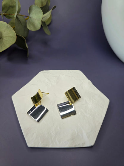 Duo-Tone Geometry drops displayed on a white textured hexagon stone. These 3.2cm mixed metal earrings feature interlocking gold and silver squares with 925 silver needles - My Silver Lining.