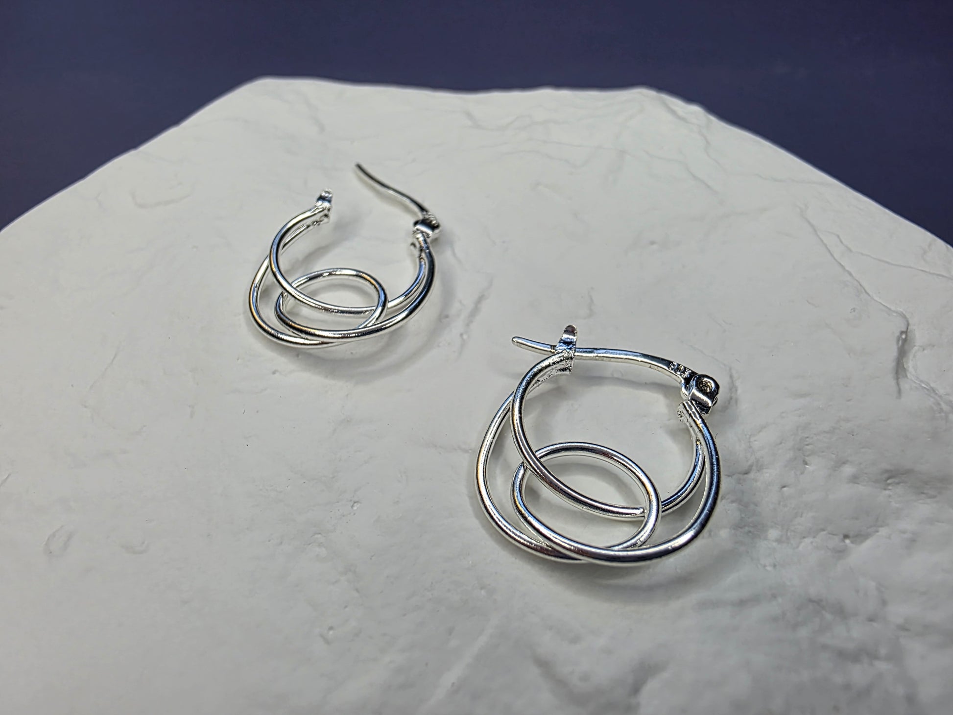 Eternal Embrace hoops displayed on a white textured hexagon stone. These S925 sterling silver interlocking double-hoop earrings offer a minimalist and dimensional look - My Silver Lining.