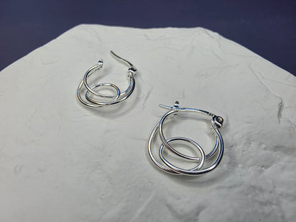 Eternal Embrace hoops displayed on a white textured hexagon stone. These S925 sterling silver interlocking double-hoop earrings offer a minimalist and dimensional look - My Silver Lining.