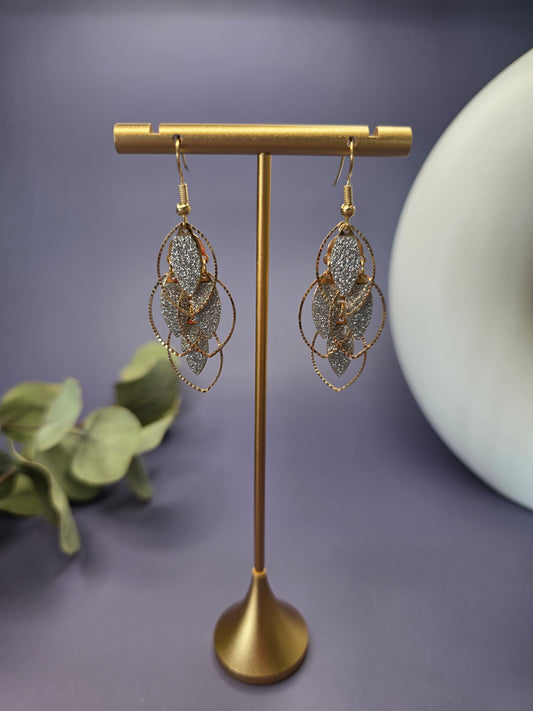 Glitzy Filigree Leaf earrings suspended from a golden T-bar jewelry stand. These 6cm 18K gold plated botanical earrings showcase a multi-tiered leaf design with a sparkling dusty texture and stainless steel needles - My Silver Lining.