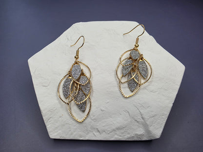 Glitzy Filigree Leaf earrings displayed on a white textured hexagon stone. These 6cm 18K gold plated botanical drop earrings feature a tiered design with a shimmering dusty texture and stainless steel ear needles - My Silver Lining.