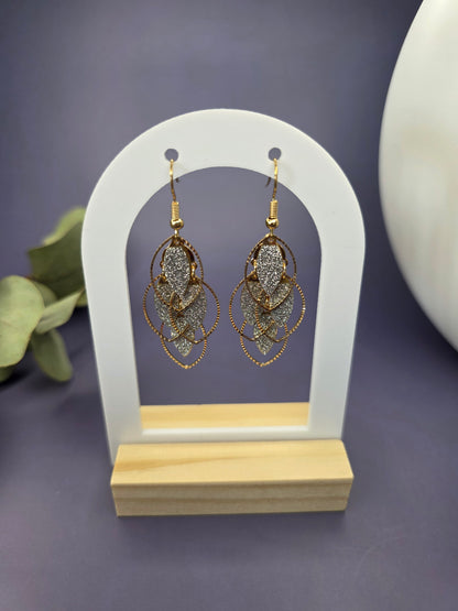 Glitzy Filigree Leaf earrings suspended from a white arch jewelry stand. These 6cm 18K gold plated botanical earrings showcase a multi-tiered leaf design with a sparkling dusty texture and stainless steel needles - My Silver Lining.