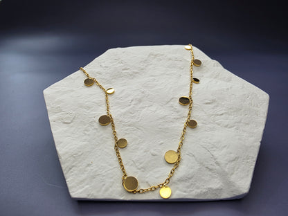 Golden Dewdrop necklace displayed on a white textured hexagon stone. This 18K gold plated 304 stainless steel necklace features dangling discs for a subtle shimmer - My Silver Lining.