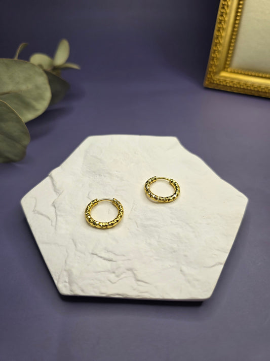 Golden ripple huggies displayed on a white textured hexagon stone. These 1.7cm KC gold plated waterproof sleeper earrings made of 304 stainless steel feature a multifaceted light-catching design - My Silver Lining.