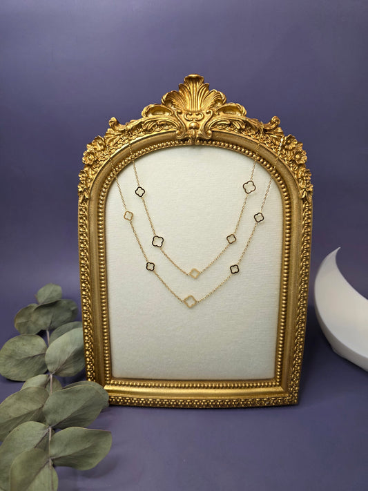 A close-up image of a long, double-layered gold-plated chain featuring alternating tiny gold eye-shaped charms and open-frame four-leaf clover charms. The necklace is displayed against a lavender background.