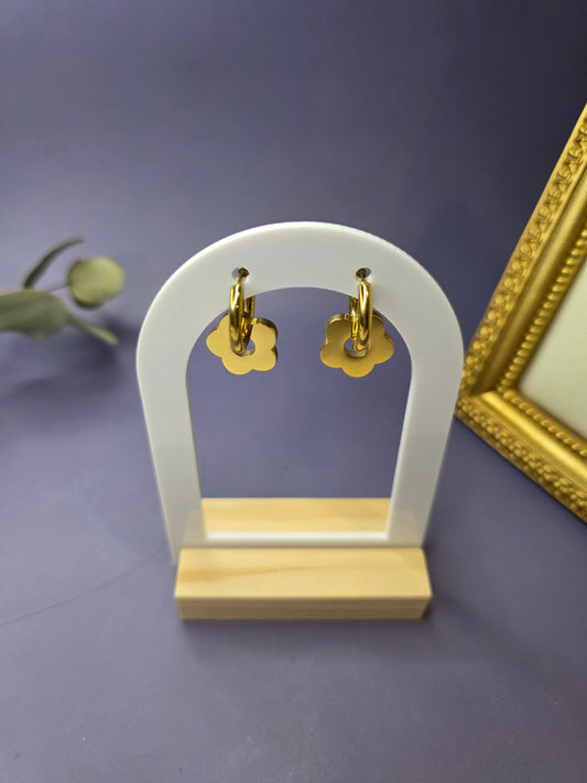 A pair of gold-toned 18K plated stainless steel hoop earrings featuring large, flat, dangling flower charms with rounded petals, set against a neutral, gold-toned fabric background.