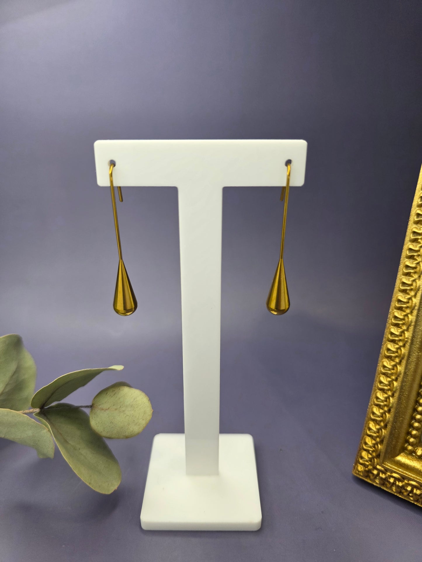 A pair of long, gold-toned 18K plated stainless steel drop earrings featuring smooth, polished teardrop-shaped pendants hanging from elongated wire hooks, displayed on a white textured surface.