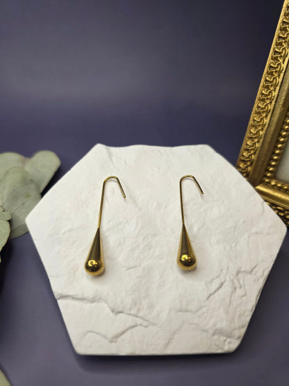 A pair of long, gold-toned 18K plated stainless steel drop earrings featuring smooth, polished teardrop-shaped pendants hanging from elongated wire hooks, displayed on a white textured surface.