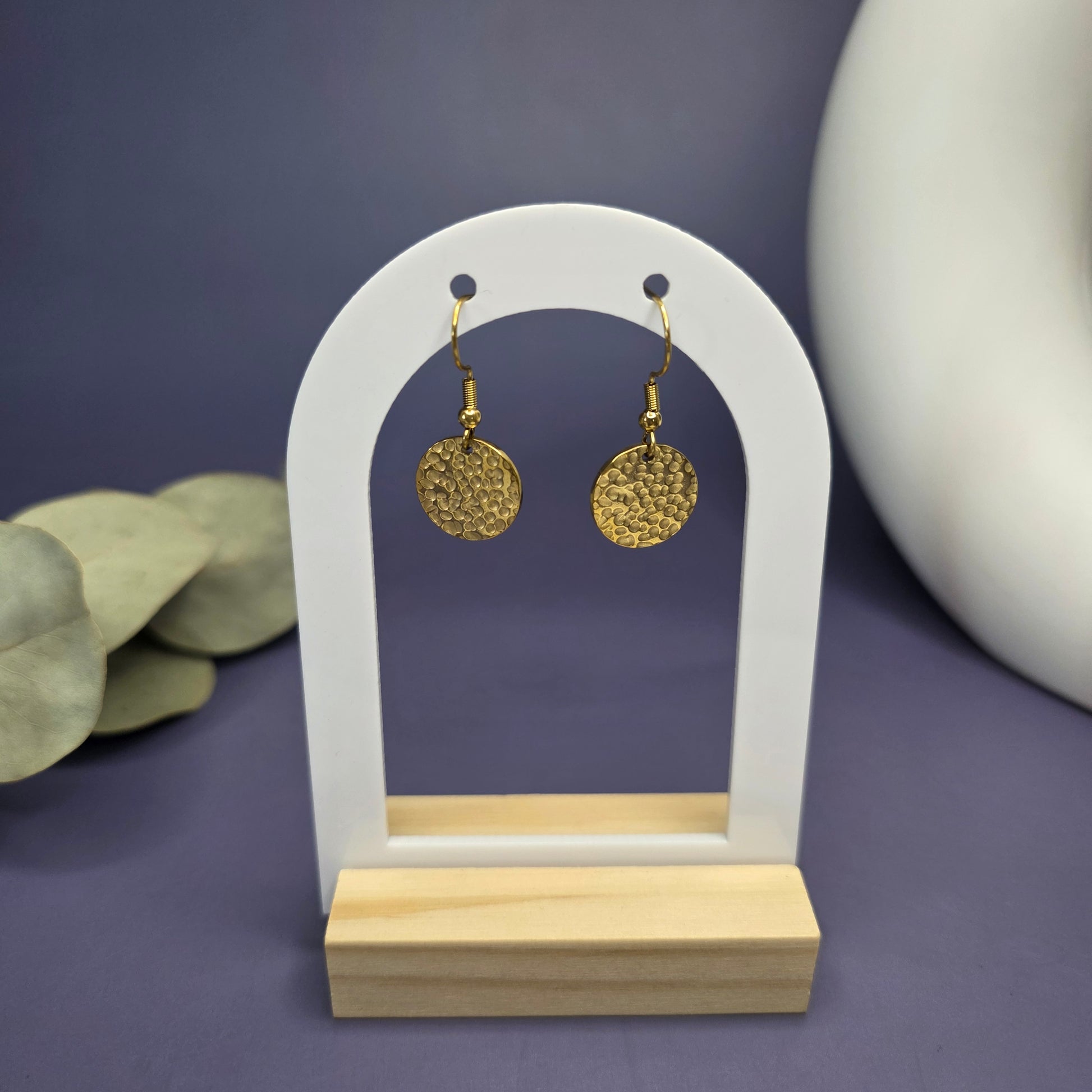 Frontal product shot of the Honey Gold Hammered Drops, displaying the pair of textured gold-toned disc earrings on a modern white and wood arch stand.

Caption/Title: Honey Gold Hammered Drops on display.