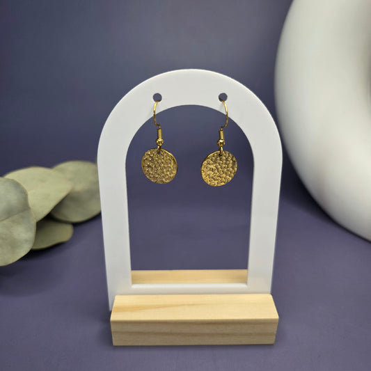 Frontal product shot of the Honey Gold Hammered Drops, displaying the pair of textured gold-toned disc earrings on a modern white and wood arch stand.

Caption/Title: Honey Gold Hammered Drops on display.