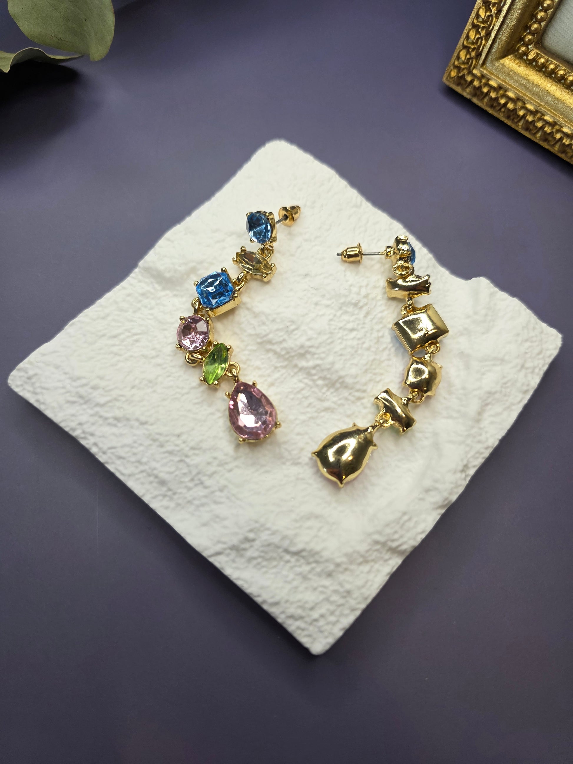 Jeweled Cascade drop earrings displayed on a white textured hexagon stone. These 7cm 18K gold plated chandelier earrings feature vibrant pink, blue, and clear rhinestones - My Silver Lining.