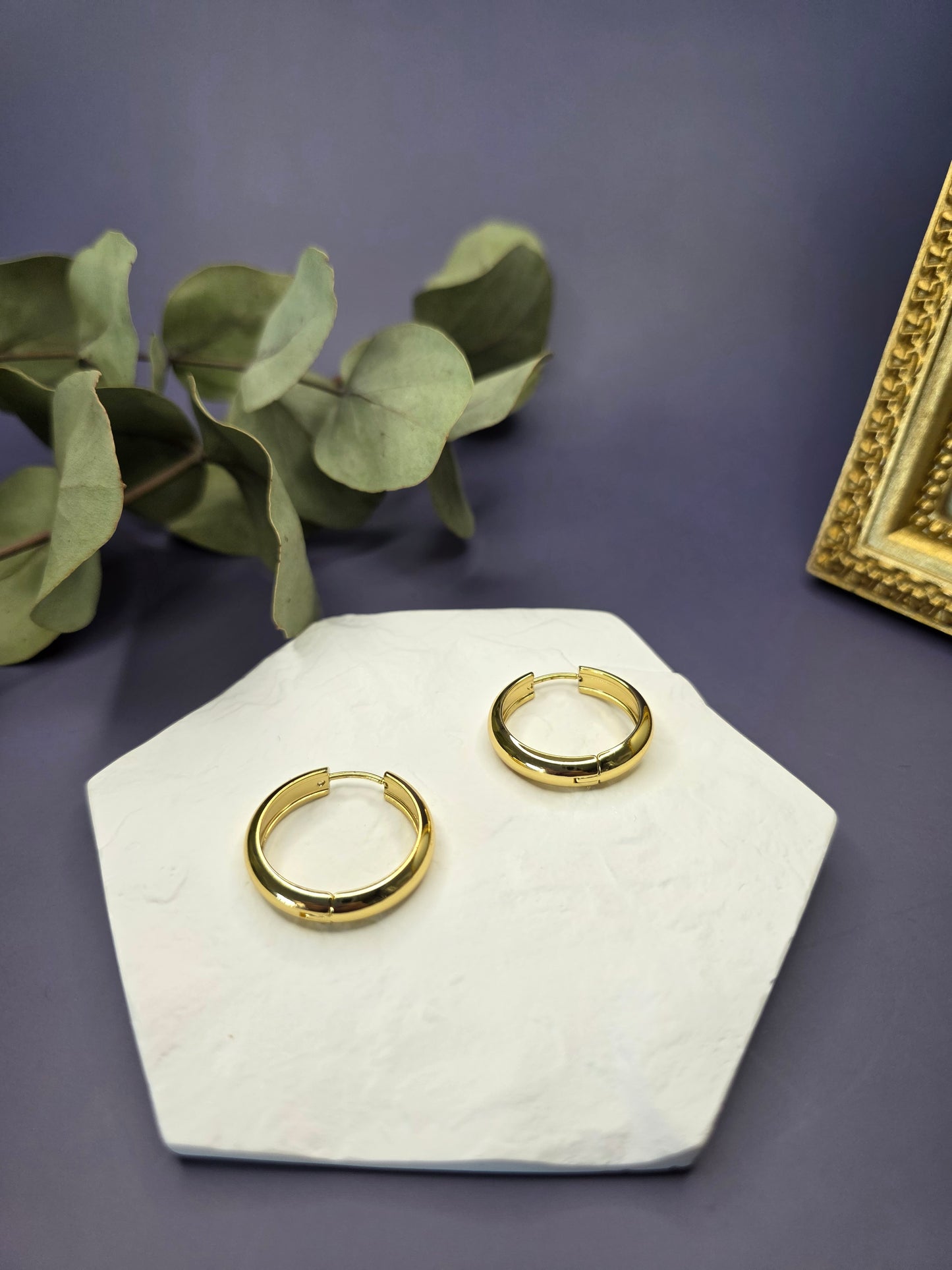 Line of Luxe hoops displayed on a white textured hexagon stone. These 2.5cm 18K gold plated waterproof earrings made of 304 stainless steel feature a wide huggie design - My Silver Lining.