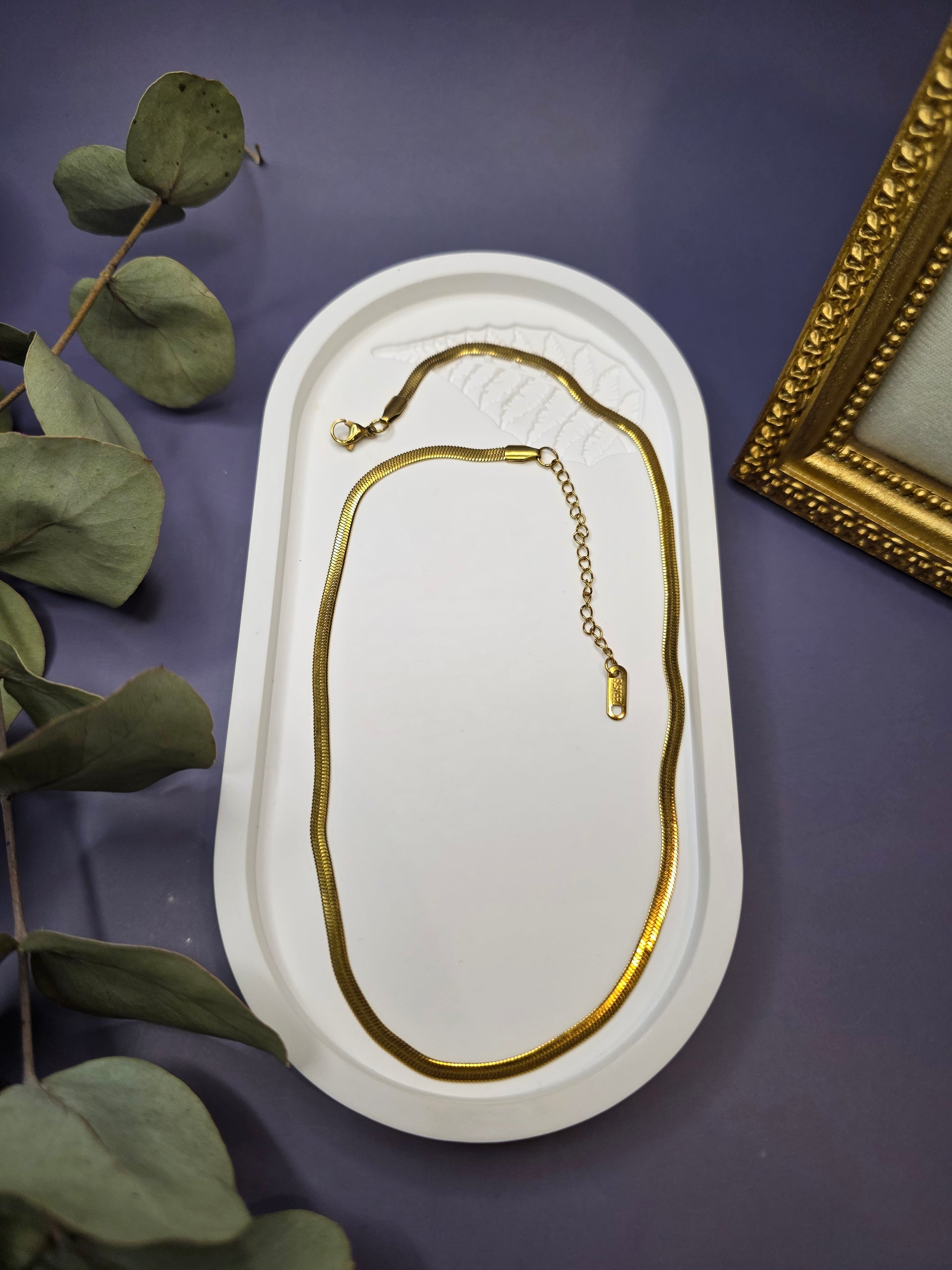 Liquid Gold Snake Chain necklace displayed in a white oval stone tray. This waterproof 18K gold plated flat snake chain offers a radiant, mirror-polished finish - My Silver Lining.