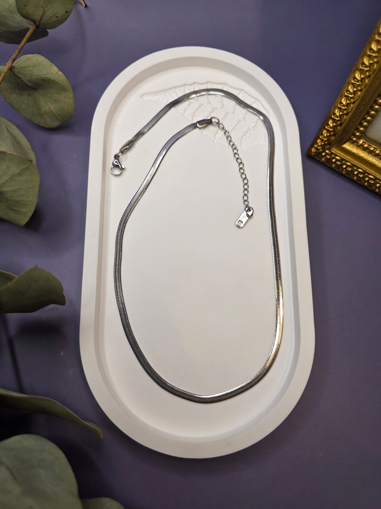Liquid Silver Snake Chain necklace displayed in a white oval stone tray. This waterproof stainless steel flat snake chain offers a radiant, mirror-polished silver finish - My Silver Lining.