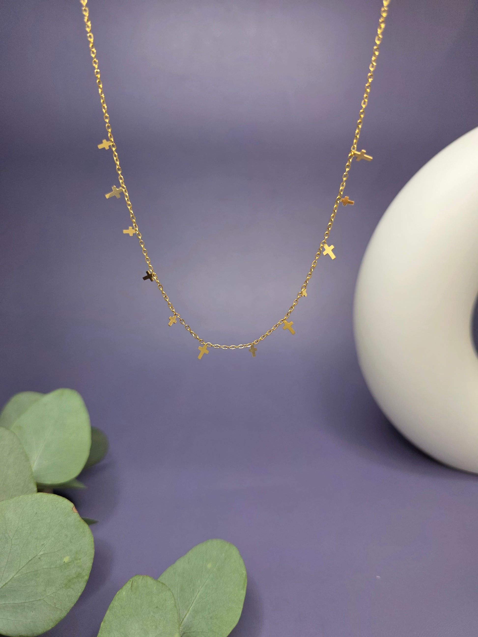 Close-up of the Mini Cross Charm Necklace in a floating composition against a purple background. This waterproof 18k gold plated jewelry features 0.4cm and 0.6cm cross charms, offering a refined and contemporary aesthetic.