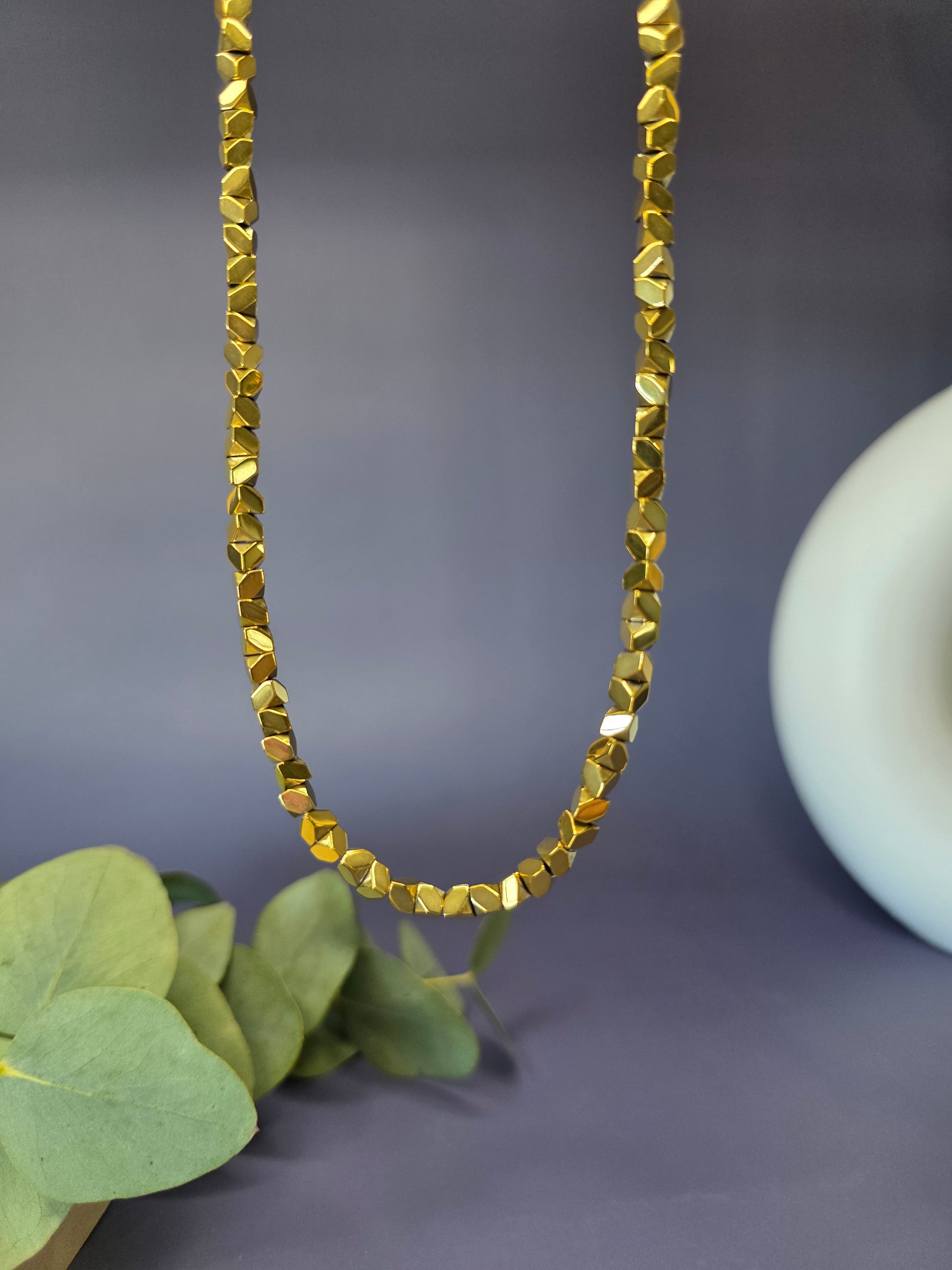 The Solaris Chain necklace, made of small, gold-toned faceted beads, displayed on a beige jewelry bust. This stainless steel chain is $40 \text{ cm}$ long with a $5 \text{ cm}$ extender, perfect for a minimalist and boho style.