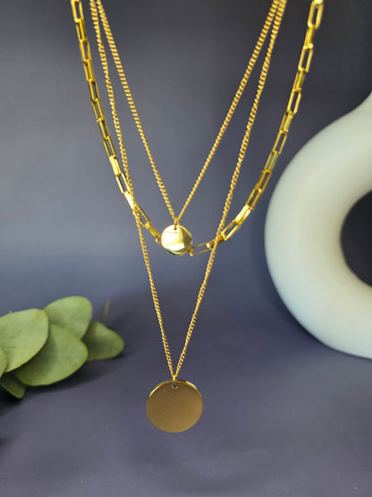 The Golden Harmony Trio, an stainless steel three-layer necklace set, displayed on a beige jewelry bust. The single-piece design includes a small disc, a rectangular link chain, and a large, reflective disc pendant, showcasing a perfectly stacked look.