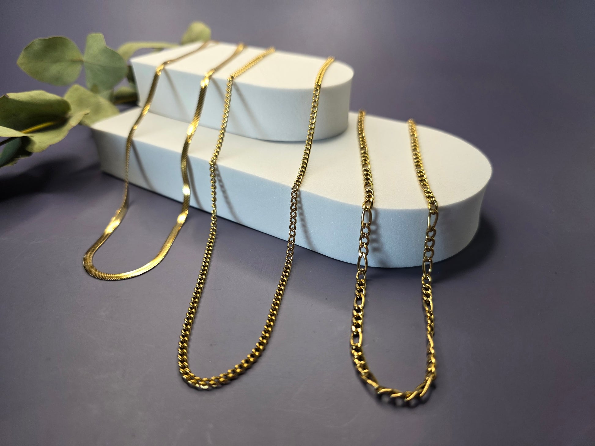 The Golden Texture Trio, a set of three separate gold-plated stainless steel necklaces for layering. The set includes a smooth herringbone chain, a classic figaro chain, and a fine twisted chain.