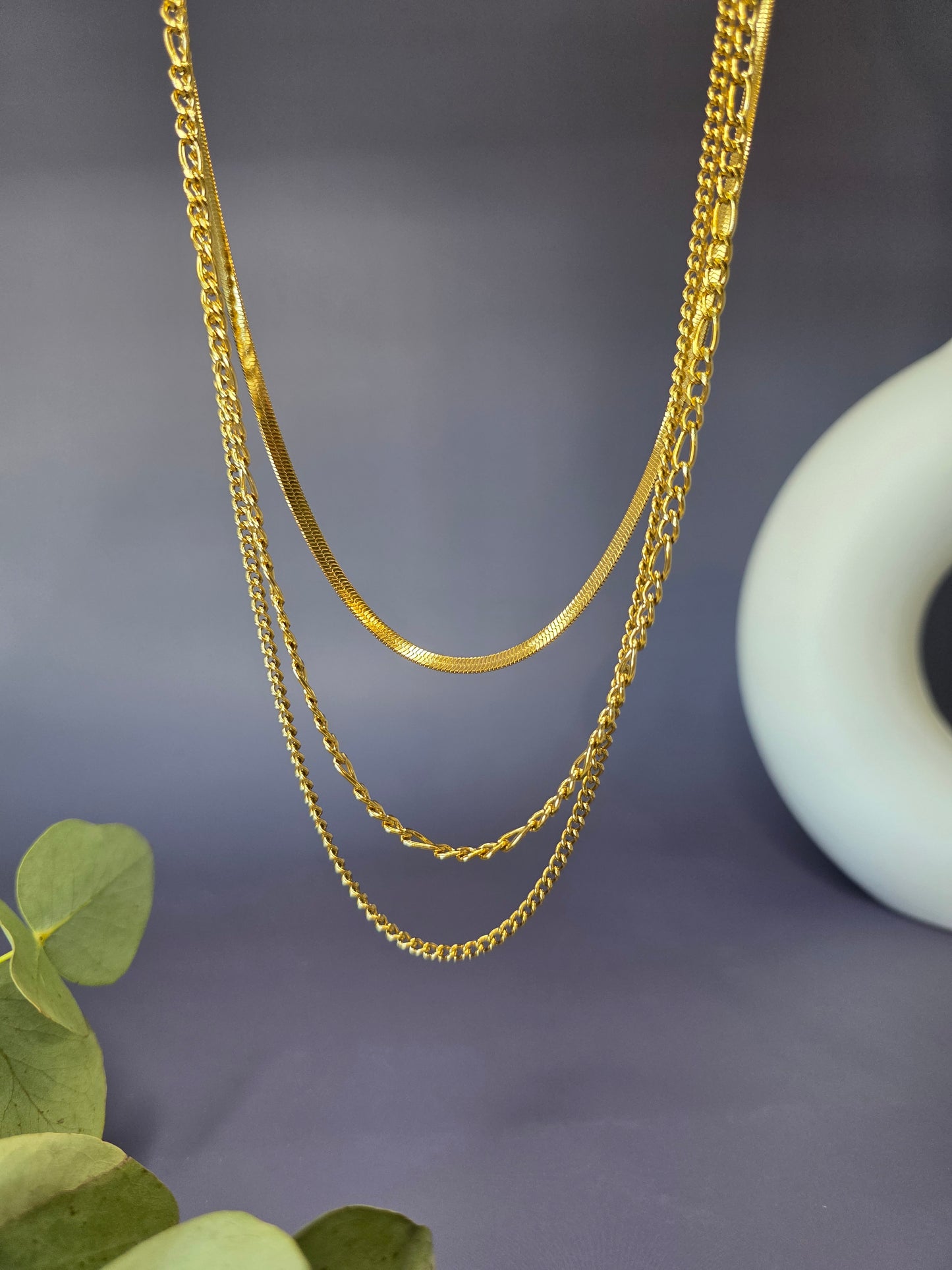 The Golden Texture Trio, a set of three separate gold-plated stainless steel necklaces for layering. The set includes a smooth herringbone chain, a classic figaro chain, and a fine twisted chain.