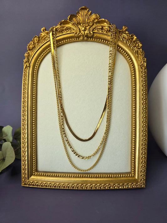 The Golden Texture Trio, a set of three separate gold-plated stainless steel necklaces for layering. The set includes a smooth herringbone chain, a classic figaro chain, and a fine twisted chain.