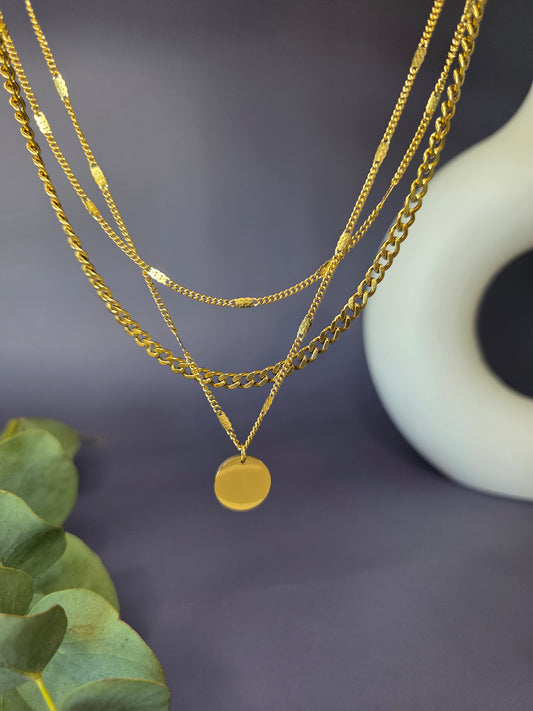 Close-up of The Luminous Trio necklace, an KC gold-plated stainless steel three-layer set. It highlights the intricate details of the delicate fine chain, the robust cuban link chain, and the beaded satellite chain with a polished disc pendant, hanging against a blurred background