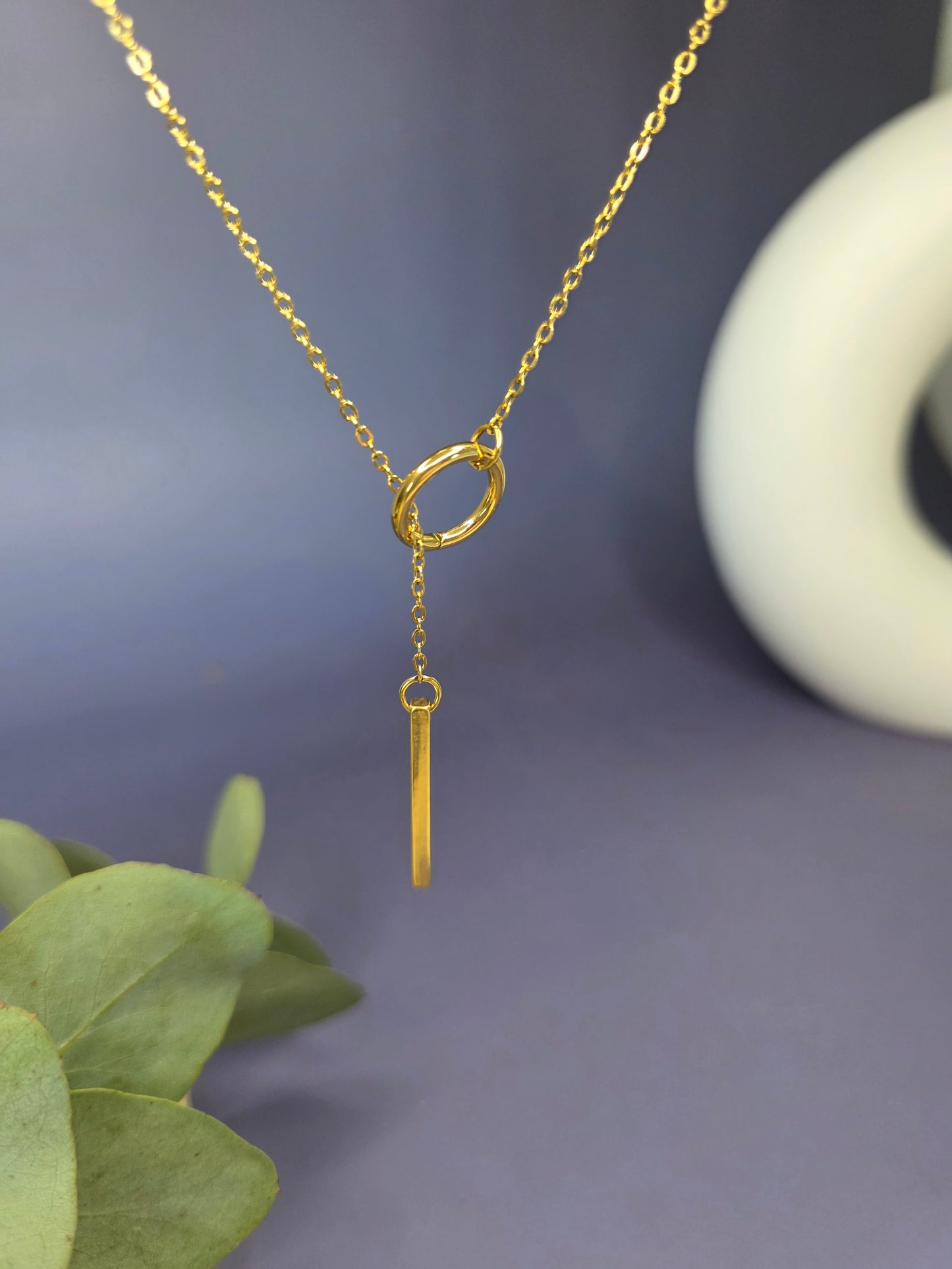 The Sleek Gold Y-Necklace Choker, a gold-plated stainless steel lariat necklace featuring a fine chain, a small circular component, and a polished vertical bar drop pendant