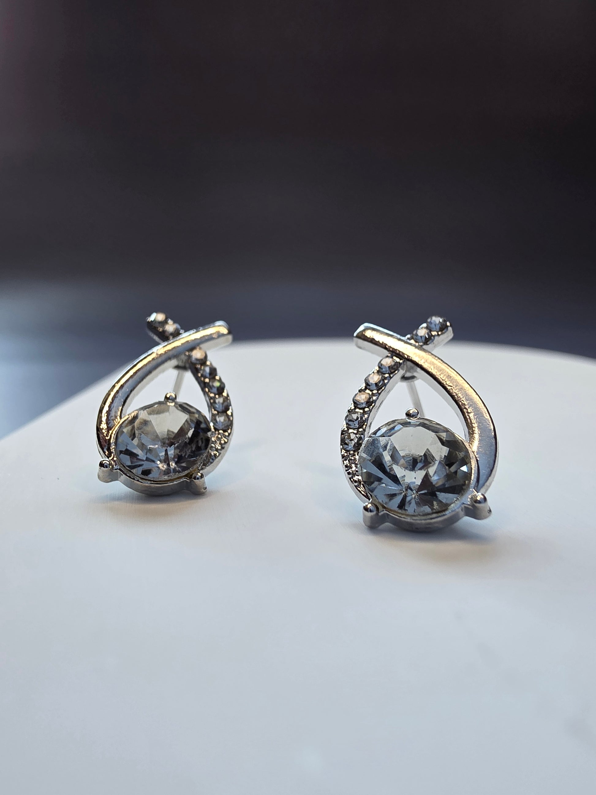 Nova Orbit earrings displayed on a white textured hexagon stone. These 1.8cm silver-toned earrings feature a large central stone and an asymmetrical rhinestone-lined frame - My Silver Lining.