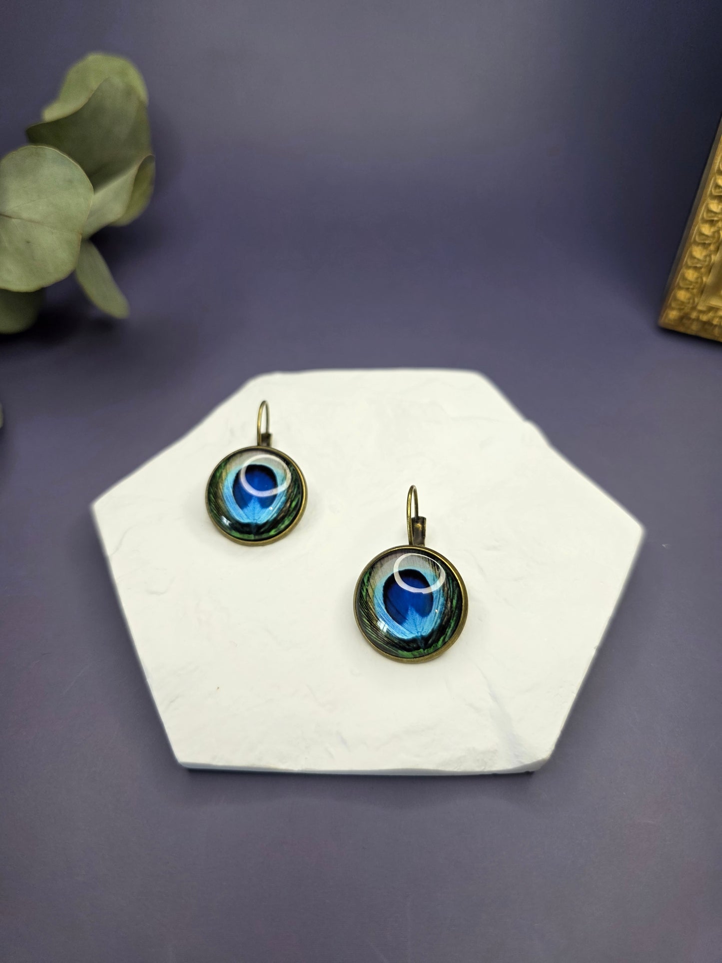 Peacock's Eye Drops displayed on a white textured hexagon stone. These 3cm bohemian glass dome earrings feature a vibrant peacock feather design in an antique frame - My Silver Lining.