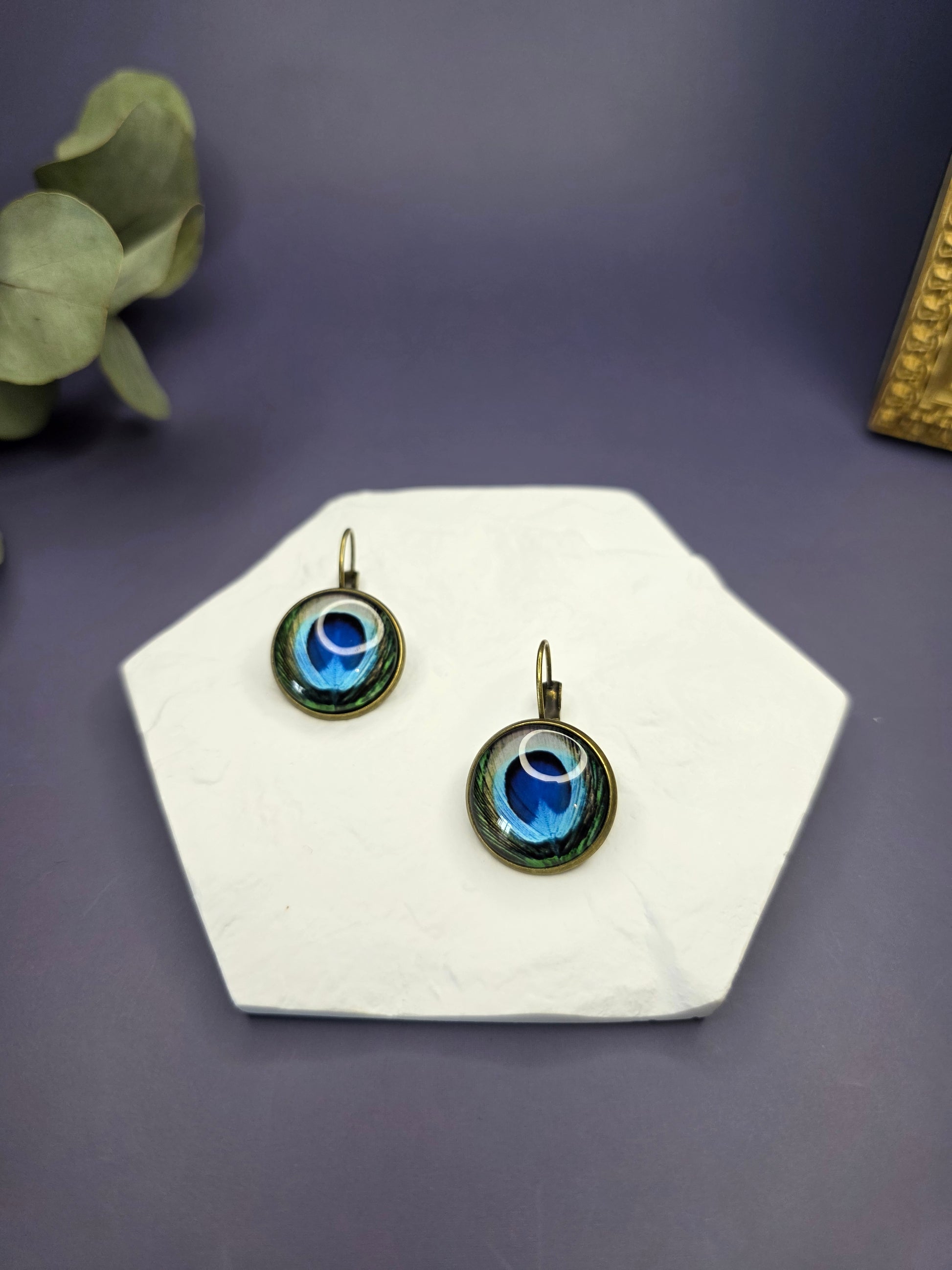 Peacock's Eye Drops displayed on a white textured hexagon stone. These 3cm bohemian glass dome earrings feature a vibrant peacock feather design in an antique frame - My Silver Lining.
