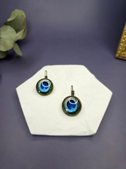Peacock's Eye Drops displayed on a white textured hexagon stone. These 3cm bohemian glass dome earrings feature a vibrant peacock feather design in an antique frame - My Silver Lining.