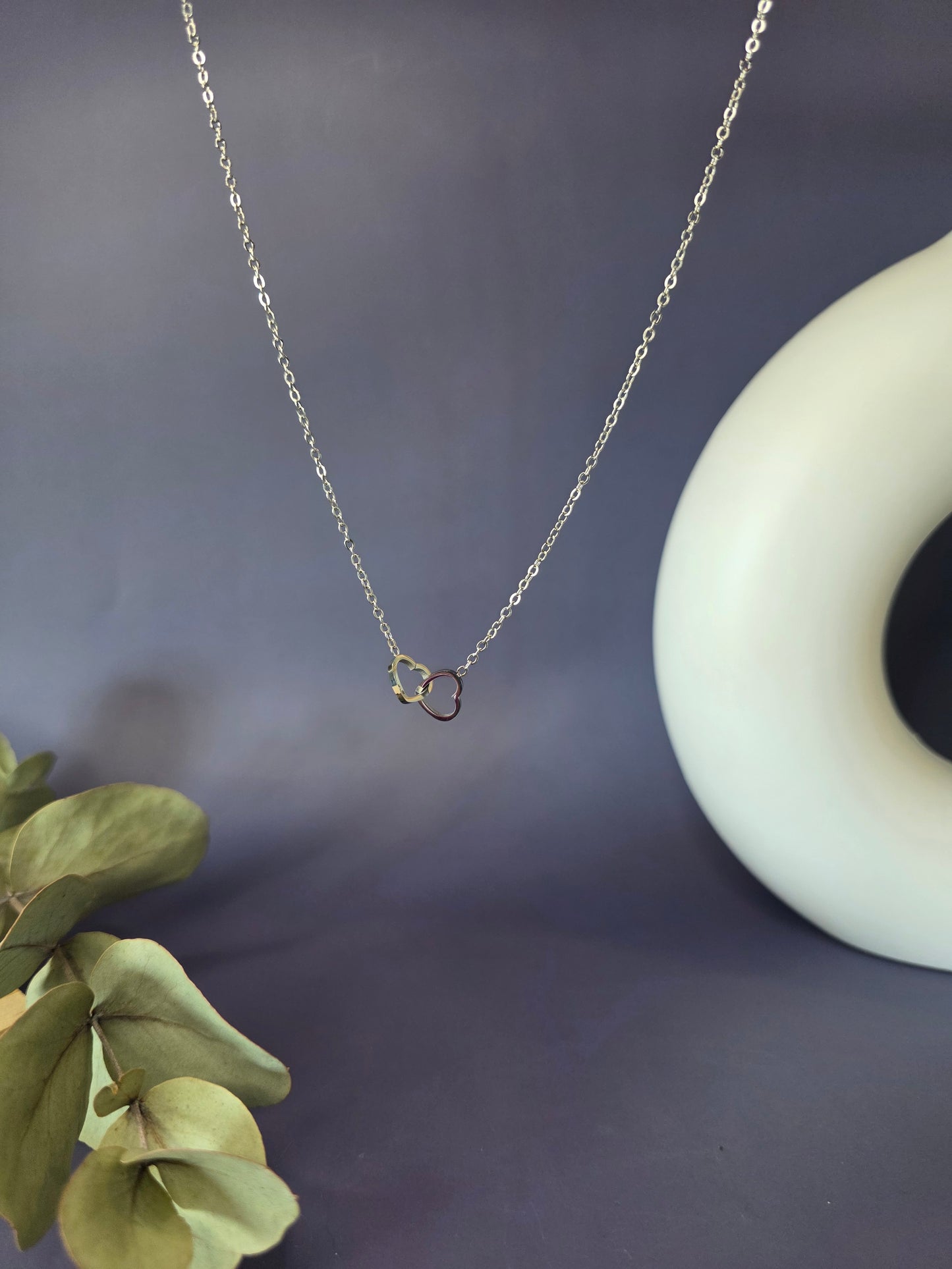Close-up of the Silver Dual Hearts necklace against a soft purple backdrop. A tarnish-proof 316 stainless steel interlocking heart pendant that perfectly embodies the message: Every day has a silver lining, wear yours - My Silver Lining.
