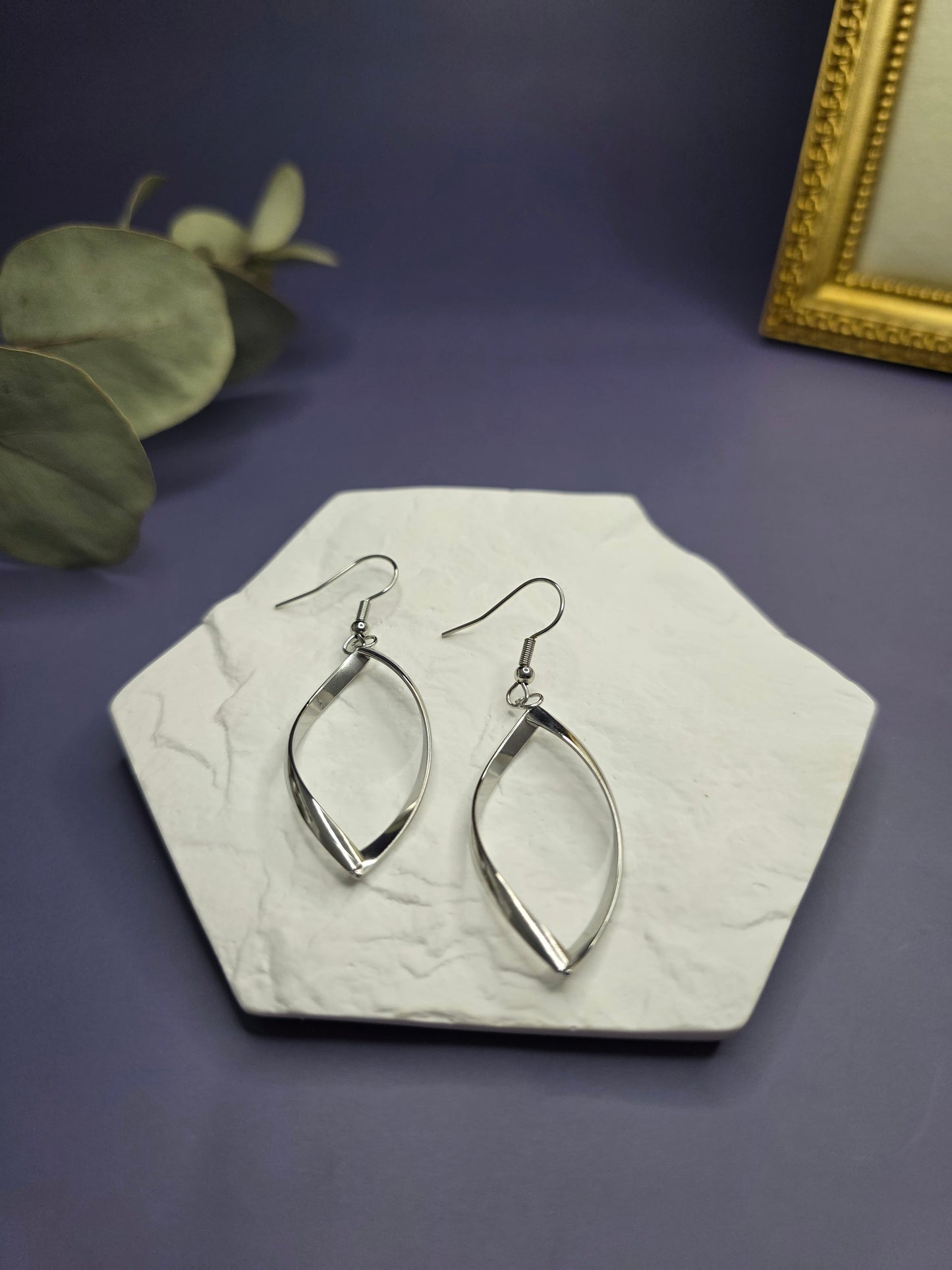 Silver ribbon earrings displayed on a white textured hexagon stone. These 7cm waterproof 316 stainless steel drop earrings offer a polished, tarnish-resistant twisted design for modern everyday elegance - My Silver Lining.