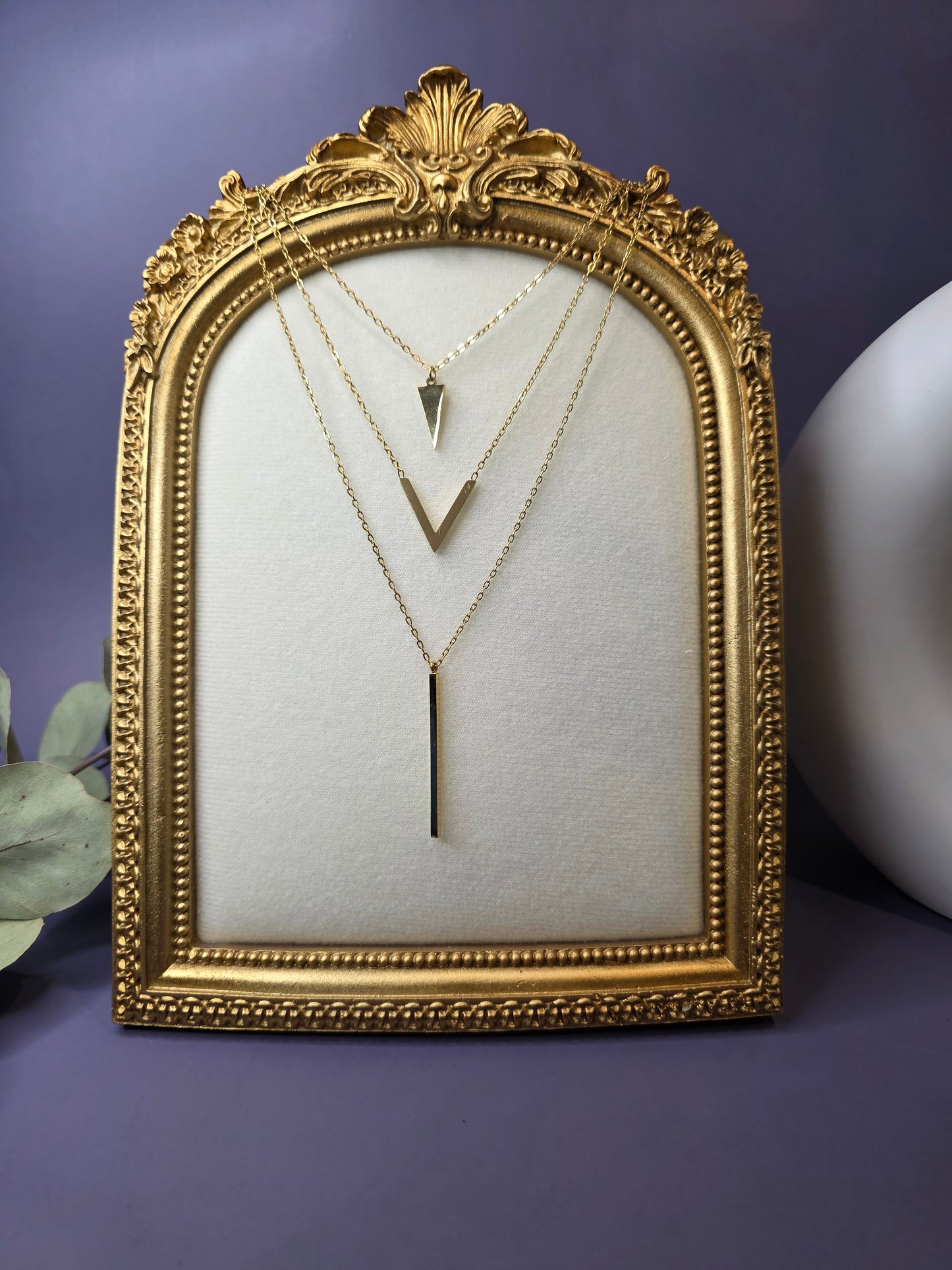Three-layered 18k gold plated stainless steel necklaces featuring a triangle, chevron V-shape, and vertical bar pendant displayed in an ornate gold vintage frame.