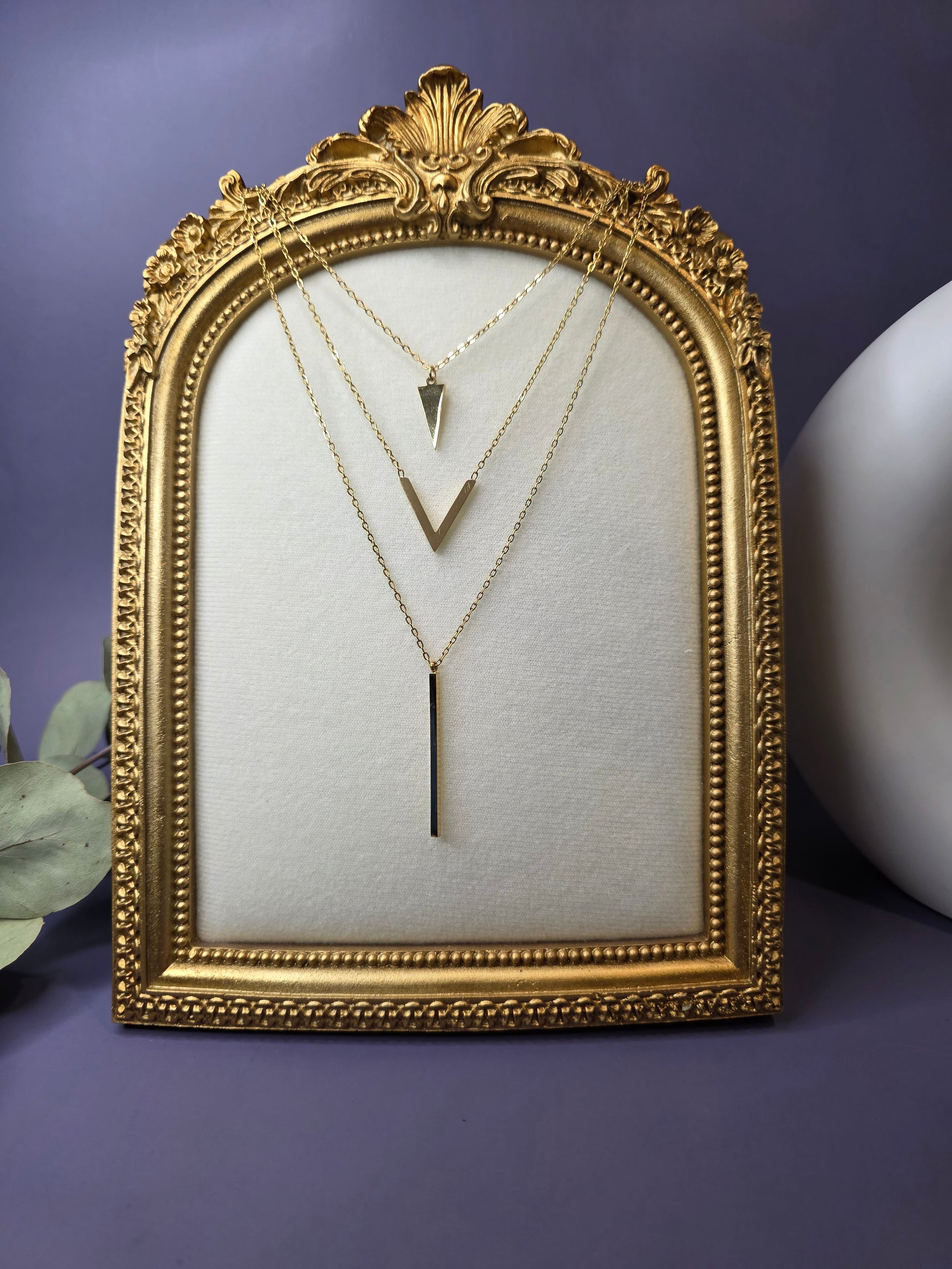 Three-layered 18k gold plated stainless steel necklaces featuring a triangle, chevron V-shape, and vertical bar pendant displayed in an ornate gold vintage frame.
